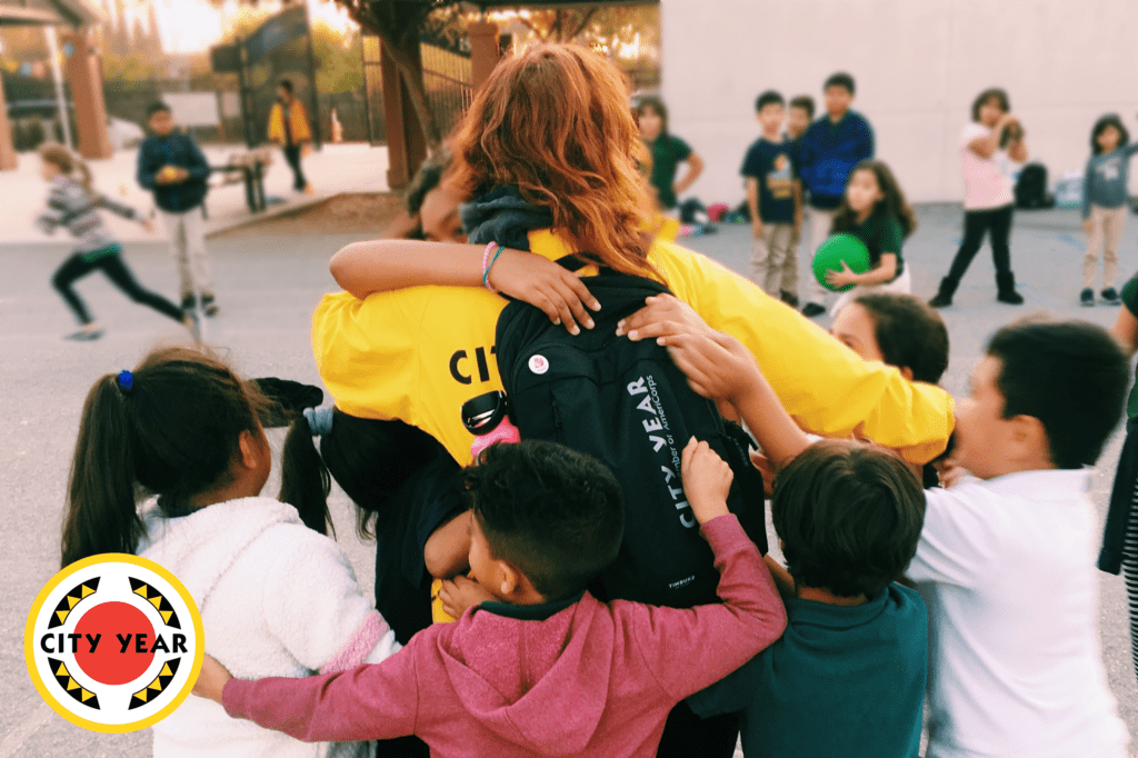 Maraina Weyl, a City Year San José AmeriCorps member, in a yellow jacket being surrounded by students at her school who are hugging her. 