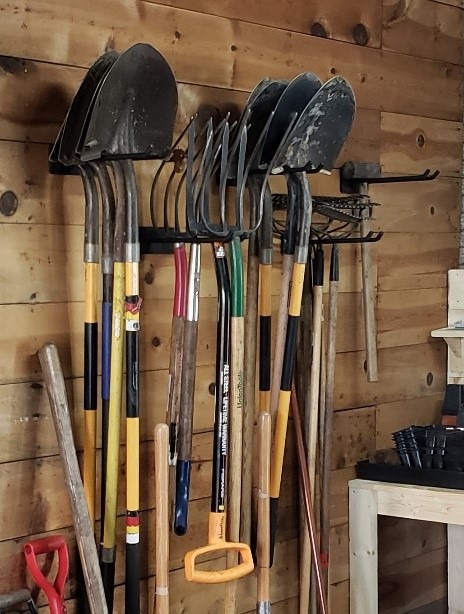 Various landscaping tools hang from two-pronged hooks against a brown wall.