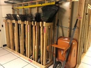 Various landscaping tools in a wooden storage rack. An orange wheelbarrow leans against the wall beside it.