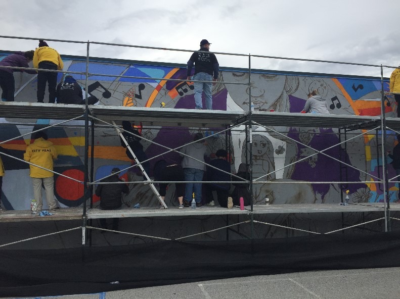 Two individuals in yellow City Year jackets and one individual in a black jacket stand on scaffolding as they work on a large mural.