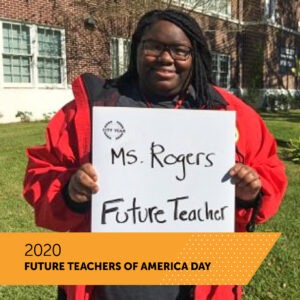 A City Year AmeriCorps member holds a board with her name Ms. Rogers and the title Future Teacher as she stands on the front lawn of her school. There is a banner that reads 2020 Future Teachers of America Day.