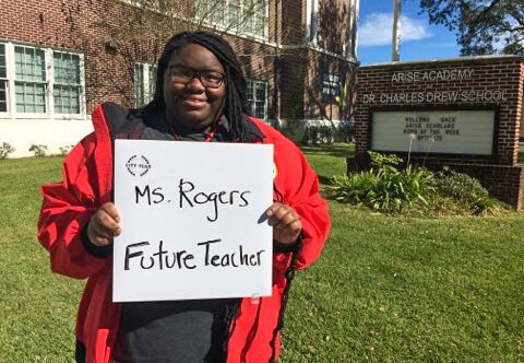 An AmeriCorps member in her red jacket smiles and holds a sign that displays her name Ms. Rogers and also says Future Teacher. She is standing on the front lawn of ARISE Academy
