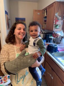 Angela, wearing an oven mitt, holds her baby, who is wearing an apron.