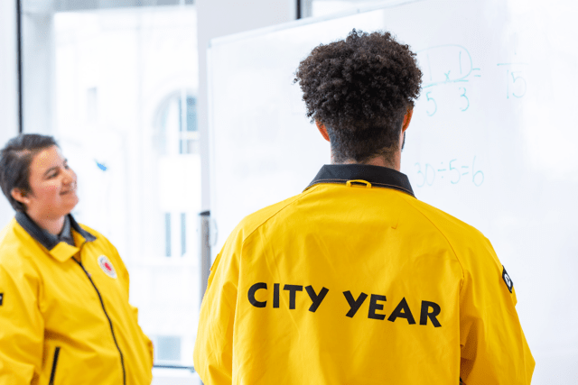 two City Year AmeriCorps members in yellow jackets face a whiteboard with math problems on it