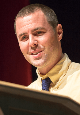 A City Year alum wearing a yellow shirt and blue tie speaks at a podium