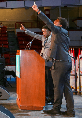 Two City Year alums in grey suits stand with an arm raised at a podium