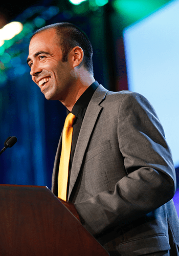 A City Year alum wearing a grey suit top and yellow tie laughs at a podium