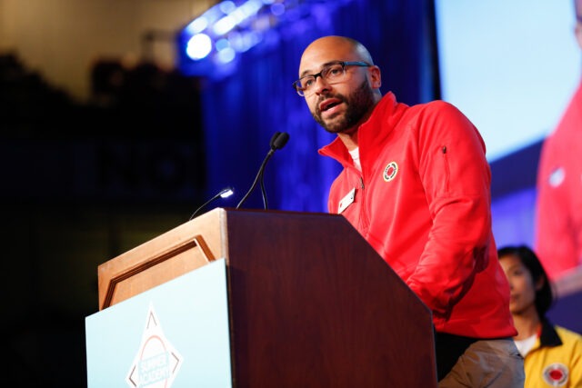A City Year alum wearing glasses and a red jacket speaks at a podium