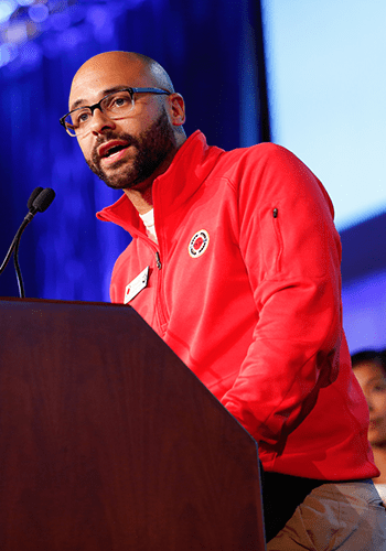 A City Year alum wearing a red City Year half-zip sweatshirt and glasses speaks at a podium