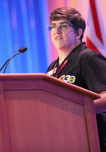 A City Year alum wearing glasses and a black City Year polo shirt stands at a podium