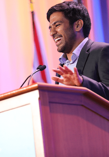 A City Year alum wearing a dark grey suit top laughs at a podium