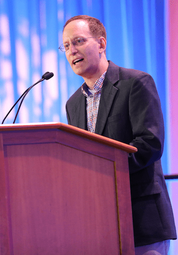 A City Year alum wearing glasses, a black suit jacket and plaid shirt speaks at a podium