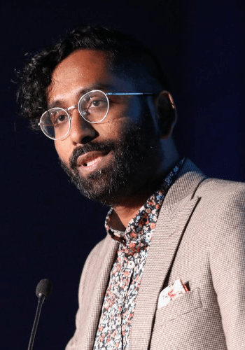 A City Year alum wearing glasses, a grey plaid suit jacket and floral shirt speaks at a podium