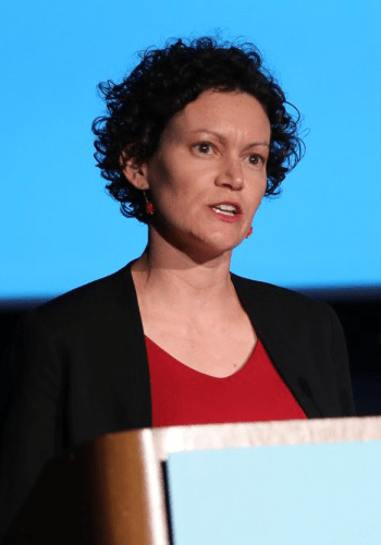 A City Year alum wearing a black suit jacket and red top speaks at a podium