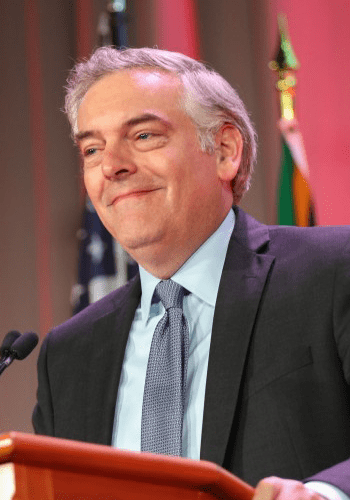 A City Year alum wearing a black suit jacket and blue tie smiles at a podium