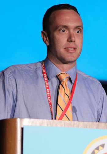 A City Year alum wearing a blue shirt and striped yellow tie stands at a podium