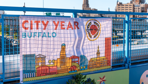 A City Year Buffalo banner with the cityscape on it hangs on a blue railing with the cityscape in the background
