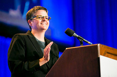 A City Year alum wearing a black blazer and glasses smiles at a podium