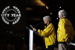 Tashia Morgridge Stands at the Podium speaking wearing Yellow City Year Jacket as husband, John, observes