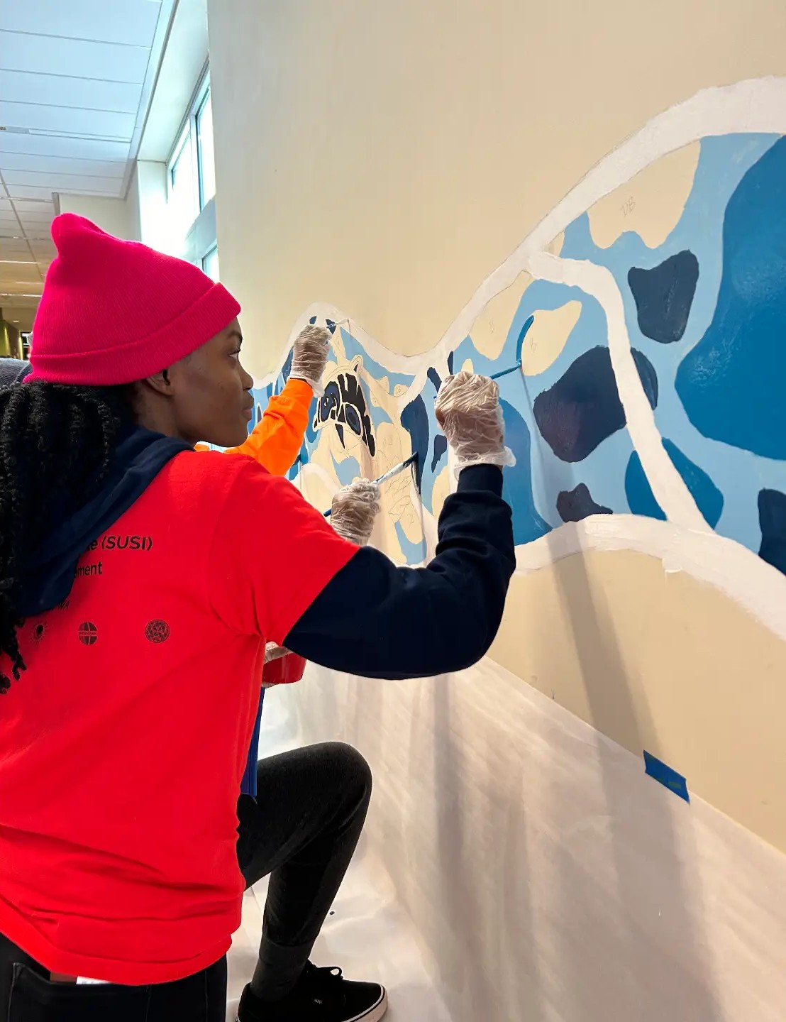 Volunteer in a red beanie painting a blue mural on a school hallway wall